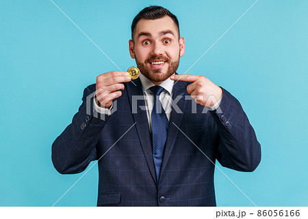 Attention to cryptocurrency. Satisfied businessman wearing official style suit pointing bitcoin and smiling to camera, showing digital money. Indoor studio shot isolated on blue background. 86056166