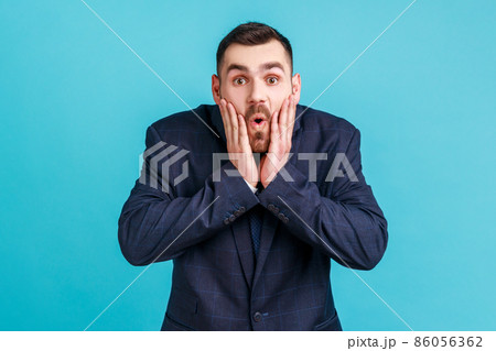 Photo of amazed bearded young adult man in official style suit looks at camera with open mouth, raising palms, being impressed of shocking news. Indoor studio shot isolated on blue background. Photo of amazed bearded young adult man in official style suit looks at camera with open mouth, raising palms, being impressed of shocking news. Indoor studio shot isolated on blue background. 86056362