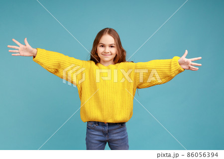 Portrait of happy cute little girl outstretching hands to embrace, giving free hugs and welcoming, wearing yellow casual style sweater. Indoor studio shot isolated on blue background. Portrait of happy cute little girl outstretching hands to embrace, giving free hugs and welcoming, wearing yellow casual style sweater. Indoor studio shot isolated on blue background. 86056394
