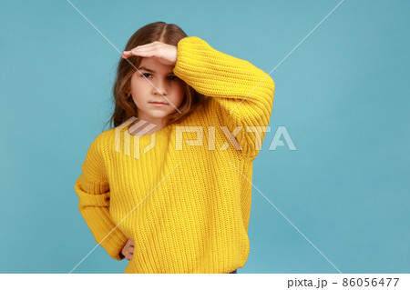 Nosy attentive serious little girl holding hand over eyes and looking far away, observing horizon, wearing yellow casual style sweater. Indoor studio shot isolated on blue background. Nosy attentive serious little girl holding hand over eyes and looking far away, observing horizon, wearing yellow casual style sweater. Indoor studio shot isolated on blue background. 86056477