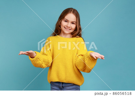 Portrait of little girl opens hands wide to give free hugs, ready to embrace friend, hospitable kind, wearing yellow casual style sweater. Indoor studio shot isolated on blue background. Portrait of little girl opens hands wide to give free hugs, ready to embrace friend, hospitable kind, wearing yellow casual style sweater. Indoor studio shot isolated on blue background. 86056518