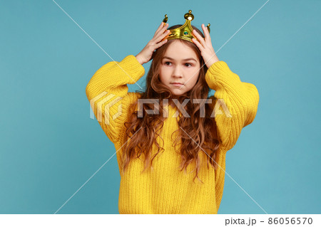 Portrait of little princess girl standing and wear golden diadem crown with smile, looking at camera, wearing yellow casual style sweater. Indoor studio shot isolated on blue background. Portrait of little princess girl standing and wear golden diadem crown with smile, looking at camera, wearing yellow casual style sweater. Indoor studio shot isolated on blue background. 86056570