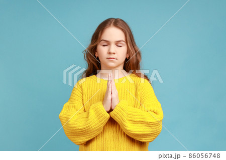 Portrait of little girl holding fingers in mudra gesture, feeling calm positive and relaxed, yoga practice, wearing yellow casual style sweater. Indoor studio shot isolated on blue background. Portrait of little girl holding fingers in mudra gesture, feeling calm positive and relaxed, yoga practice, wearing yellow casual style sweater. Indoor studio shot isolated on blue background. 86056748