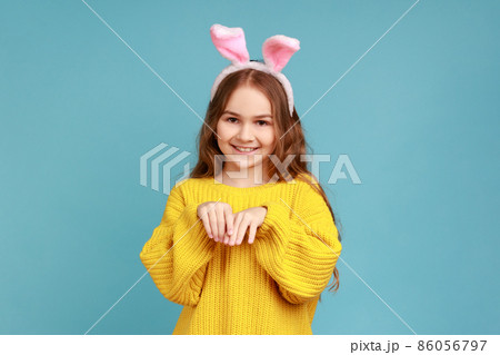 Portrait of little girl stands with pink bunny ears, child having fun playing game, looks at camera, wearing yellow casual style sweater. Indoor studio shot isolated on blue background. Portrait of little girl stands with pink bunny ears, child having fun playing game, looks at camera, wearing yellow casual style sweater. Indoor studio shot isolated on blue background. 86056797