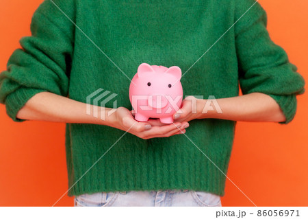 Unknown woman wearing green casual style sweater standing holding in hands piggy bank, investment, saving money, currency, deposit. Indoor studio shot isolated on orange background. 86056971