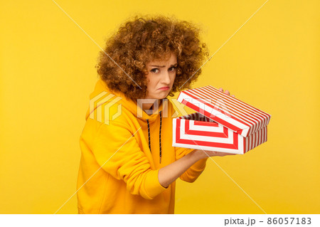 Displeased unhappy woman with curly hair unpacking gift box and looking at camera with frustrated dissatisfied expression, wearing casual style hoodie. Indoor studio shot isolated on yellow background Displeased unhappy woman with curly hair unpacking gift box and looking at camera with frustrated dissatisfied expression, wearing casual style hoodie. Indoor studio shot isolated on yellow background 86057183
