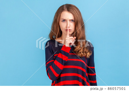 Keep silence and be quiet. Woman wearing striped casual style sweater looking angrily and holding finger on lips, gesturing shut up, stop talking. Indoor studio shot isolated on blue background. 86057501