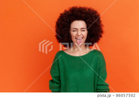 Positive playful woman with Afro hairstyle wearing green sweater standing with closed eyes and tongue out, demonstrating childish behavior. Indoor studio shot isolated on orange background. Positive playful woman with Afro hairstyle wearing green sweater standing with closed eyes and tongue out, demonstrating childish behavior. Indoor studio shot isolated on orange background. 86057502