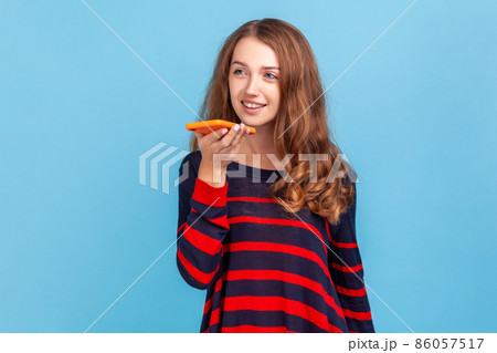 Happy young adult woman wearing striped casual style sweater, standing with smart phone, recording voice message with positive expression. Indoor studio shot isolated on blue background. 86057517