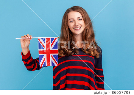Attractive woman wearing striped casual style sweater, holding flag of a constituent unit of the United Kingdom, celebrating British Independence Day Indoor studio shot isolated on blue background. 86057703