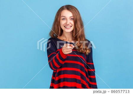 Hey you. Happy woman wearing striped casual style sweater pointing to camera and looking with charming toothy smile, indicating finger, making choice. Indoor studio shot isolated on blue background. Hey you. Happy woman wearing striped casual style sweater pointing to camera and looking with charming toothy smile, indicating finger, making choice. Indoor studio shot isolated on blue background. 86057713