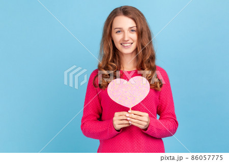 Romantic woman wearing pink pullover, holding heart symbol on stick and smiling to camera, congratulating on Valentines Day, saying I love you. Indoor studio shot isolated on blue background. Romantic woman wearing pink pullover, holding heart symbol on stick and smiling to camera, congratulating on Valentines Day, saying I love you. Indoor studio shot isolated on blue background. 86057775