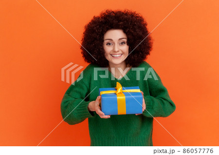 Happy positive smiling woman with Afro hairstyle wearing green casual style sweater holding wrapped gift box, looking at camera, congratulating. Indoor studio shot isolated on orange background. Happy positive smiling woman with Afro hairstyle wearing green casual style sweater holding wrapped gift box, looking at camera, congratulating. Indoor studio shot isolated on orange background. 86057776