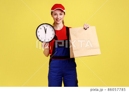 Portrait of happy positive courier woman holding parcel and wall clock, fast delivery service, delivering in time, wearing overalls and red cap. Indoor studio shot isolated on yellow background. Portrait of happy positive courier woman holding parcel and wall clock, fast delivery service, delivering in time, wearing overalls and red cap. Indoor studio shot isolated on yellow background. 86057785