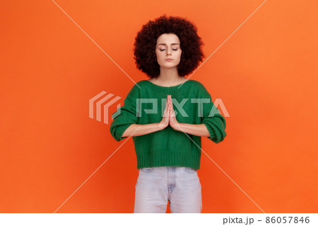 Calm relaxed young adult woman with Afro hairstyle wearing green casual style sweater doing yoga exercise breath technique reduce stress. Indoor studio shot isolated on orange background. 86057846