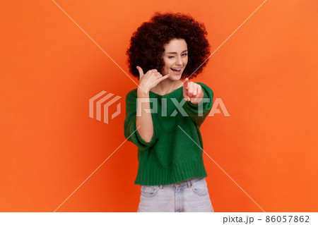 Hey you, call me. Woman with Afro hairstyle standing with telephone hand gesture and smiling to camera, flirting offering to contact by phone. Indoor studio shot isolated on orange background. Hey you, call me. Woman with Afro hairstyle standing with telephone hand gesture and smiling to camera, flirting offering to contact by phone. Indoor studio shot isolated on orange background. 86057862