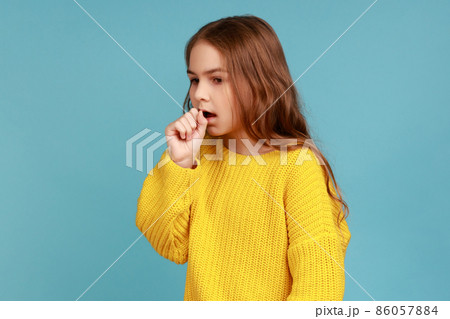 Portrait of unhealthy sick little girl having flu symptoms, coughing, keeping hand near mouth, wearing yellow casual style sweater. Indoor studio shot isolated on blue background. Portrait of unhealthy sick little girl having flu symptoms, coughing, keeping hand near mouth, wearing yellow casual style sweater. Indoor studio shot isolated on blue background. 86057884