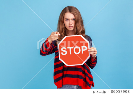 Serious woman wearing striped casual style sweater, holding big red stop road sign and pointing finger on you, strictly looking at camera. Indoor studio shot isolated on blue background. 86057908