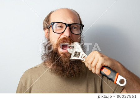 Portrait of male trying to pull out bad tooth with adjustable wrench , keeps mouth opened, bald bearded man wearing T-shirt and glasses. Indoor studio shot isolated on gray background. Portrait of male trying to pull out bad tooth with adjustable wrench , keeps mouth opened, bald bearded man wearing T-shirt and glasses. Indoor studio shot isolated on gray background. 86057961