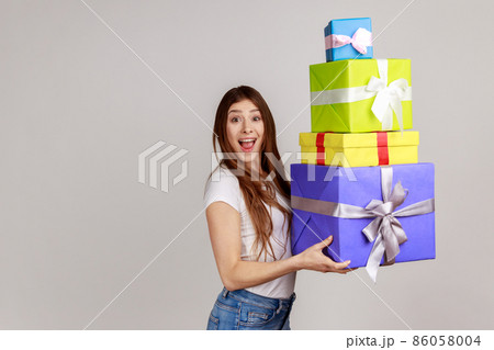 Surprised excited woman holding lot of wrapped boxes and looking with amazement, shocked by mount of bonuses and gifts, wearing white T-shirt. Indoor studio shot isolated on gray background. 86058004