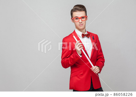 Portrait of handsome elegant gentleman with trendy glasses and stylish hairdo in red tuxedo holding big burning candle and looking at camera with alluring eyes, romantic atmosphere. indoor studio shot Portrait of handsome elegant gentleman with trendy glasses and stylish hairdo in red tuxedo holding big burning candle and looking at camera with alluring eyes, romantic atmosphere. indoor studio shot 86058226
