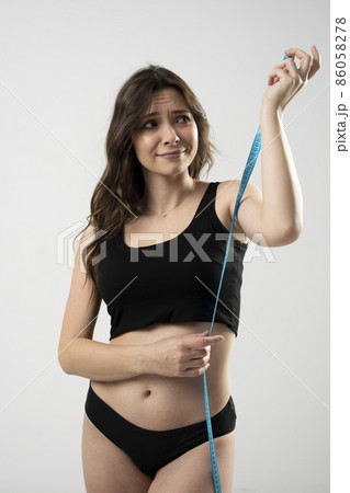 Smiling beautiful confused young brunette woman 20s wearing black casual top and hold blue centimeter tape, measure tape isolated on white wall background studio. Smiling beautiful confused young brunette woman 20s wearing black casual top and hold blue centimeter tape, measure tape isolated on white wall background studio. 86058278