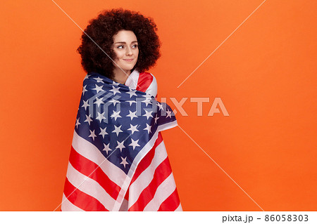 Calm dreamy woman with Afro hairstyle wearing green casual style sweater standing wrapped in USA flag, looking away, human rights. Indoor studio shot isolated on orange background. Calm dreamy woman with Afro hairstyle wearing green casual style sweater standing wrapped in USA flag, looking away, human rights. Indoor studio shot isolated on orange background. 86058303