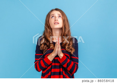 Portrait of young attractive woman wearing striped casual style sweater looking up with imploring desperate grimace, praying to god asking for help. Indoor studio shot isolated on blue background. 86058418