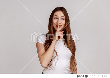 Shh, it's big secret. Beautiful cunning woman smiling, showing gesture secret sign with finger near her lips, wearing white T-shirt. Indoor studio shot isolated on gray background. 86058459
