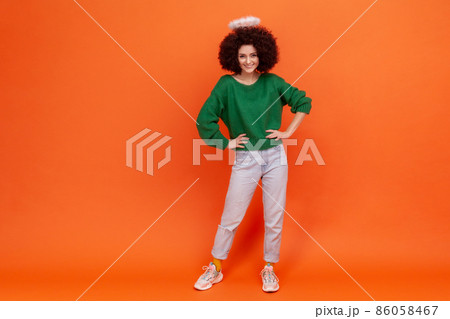 Full length of confident angelic woman with Afro hairstyle wearing green casual style sweater standing with hands on hips, having nimb over her head. Indoor studio shot isolated on orange background. Full length of confident angelic woman with Afro hairstyle wearing green casual style sweater standing with hands on hips, having nimb over her head. Indoor studio shot isolated on orange background. 86058467