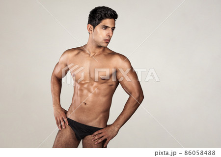 Portrait of handsome man with tanned athletic body, demonstrating his perfect sporty body, looking away, keeps hands on hips, wearing black panties. Indoor studio shot isolated on gray background. 86058488