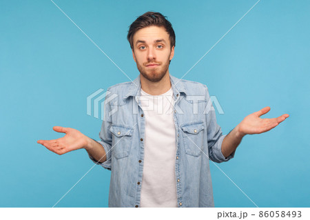 Portrait of handsome young man wearing denim shirt, shrugging shoulders, uncertain, has no answer, looking at camera with confused face. Indoor studio shot isolated on blue background. Portrait of handsome young man wearing denim shirt, shrugging shoulders, uncertain, has no answer, looking at camera with confused face. Indoor studio shot isolated on blue background. 86058493