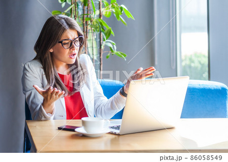 Unhappy woman employee in glasses raising hands in indignant gesture, asking why and looking angrily, looking at laptop screen, talking on video call. Indoor shot, cafe or office background. Unhappy woman employee in glasses raising hands in indignant gesture, asking why and looking angrily, looking at laptop screen, talking on video call. Indoor shot, cafe or office background. 86058549