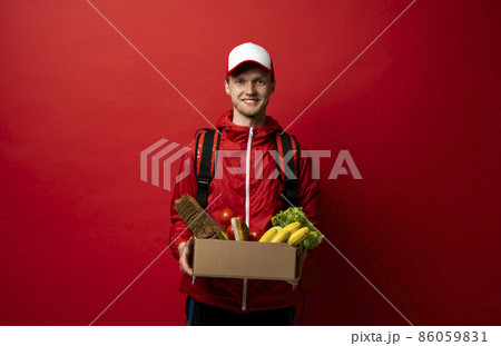 Delivery man in red uniform holds cardboard paper box with food isolated on white background, studio portrait. Service concept. Delivery man in red uniform holds cardboard paper box with food isolated on white background, studio portrait. Service concept. 86059831