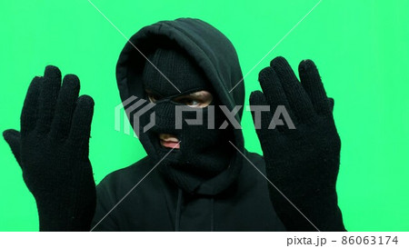 Angry young man has become a thief and prepares his equipment and clothes on a green background in a balaclava for the first store robbery Angry young man has become a thief and prepares his equipment and clothes on a green background in a balaclava for the first store robbery 86063174