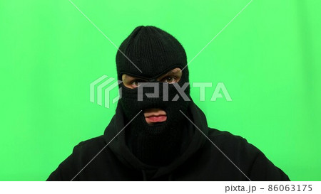 Angry young man has become a thief and prepares his equipment and clothes on a green background in a balaclava for the first store robbery Angry young man has become a thief and prepares his equipment and clothes on a green background in a balaclava for the first store robbery 86063175