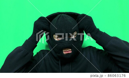 Angry young man has become a thief and prepares his equipment and clothes on a green background in a balaclava for the first store robbery Angry young man has become a thief and prepares his equipment and clothes on a green background in a balaclava for the first store robbery 86063176