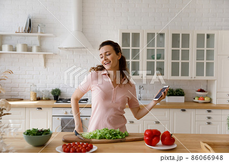 Happy carefree young woman dancing to music, cooking in kitchen. 86066948