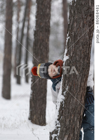 Funny child boy hides behind tree in winter forest. Boy is dressed in warm hat and winter jumpsuit. Vertical frame Funny child boy hides behind tree in winter forest. Boy is dressed in warm hat and winter jumpsuit. Vertical frame 86068481
