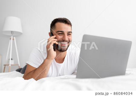 Happy young attractive guy with beard lies on white bed call by phone, looks at laptop, working or chatting Happy young attractive guy with beard lies on white bed call by phone, looks at laptop, working or chatting 86084034