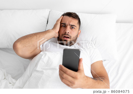 Shocked upset millennial attractive guy with beard reads message on phone and presses his hand to face Shocked upset millennial attractive guy with beard reads message on phone and presses his hand to face 86084075