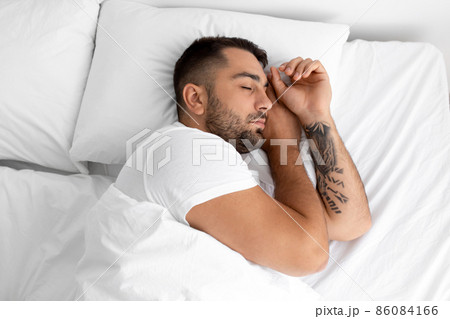Calm millennial handsome guy with beard sleeps on white comfortable bed on soft pillow in bedroom, top view Calm millennial handsome guy with beard sleeps on white comfortable bed on soft pillow in bedroom, top view 86084166