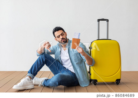 Bad travel agency service. Young arab man holding passport with tickets, showing dislike gesture, sitting near suitcase Bad travel agency service. Young arab man holding passport with tickets, showing dislike gesture, sitting near suitcase 86084689