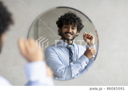 Happy bearded indian businessman standing in bathroom in front of mirror and buttoning up shirt Happy bearded indian businessman standing in bathroom in front of mirror and buttoning up shirt 86085091