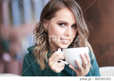 Close-up portrait of an optimistic beautiful woman with a beautiful manicure, holding a cup, sitting in a cafe, the girl looks directly into the camera. High quality photo 86090302