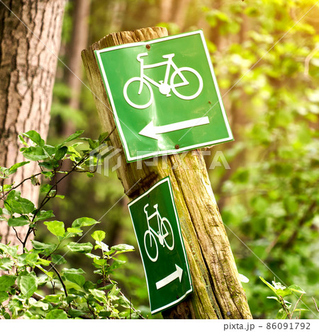 Signs for bicycle paths through a forest, green signs with white bicycles and a directional arrow on a crooked weathered wooden post 86091792