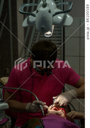 The boy is sitting in a dentist's chair, treating caries in a child, the dentist removes caries from the tooth with a drill. The boy is sitting in a dentist's chair, treating caries in a child, the dentist removes caries from the tooth with a drill. 86100589