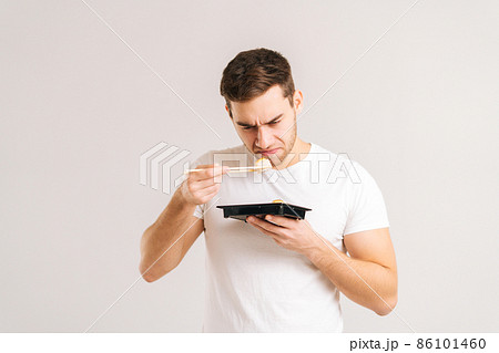 Portrait of disgusted young man holding sushi with chopsticks and sniffs with look of revulsion on face on white isolated background. Sad Caucasian guy dissatisfied with bad quality Asian food. 86101460