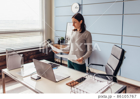 Woman standing by the desktop with documents in her hands Woman standing by the desktop with documents in her hands 86105767