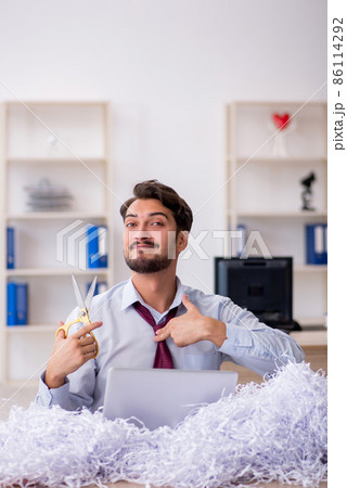 Young male employee and a lot of cut papers in the office Young male employee and a lot of cut papers in the office 86114292